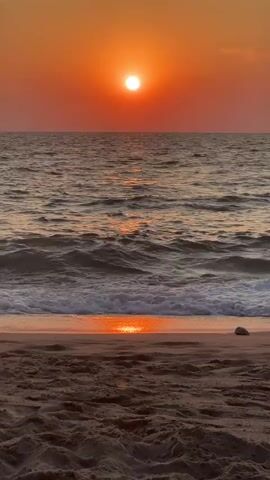 A vertical video captures a sunset over a beach. The sun, a bright white circle, is centered in the frame, low on the horizon, casting an orange glow across the sky. The ocean stretches out before the viewer, with small waves rolling towards the shore. The water reflects the sunset's colors, creating a shimmering path. The beach in the foreground is composed of sand, with a few small pebbles visible. A single, larger white pebble rests on the sand to the right of the frame. The scene is serene and appears to be uninhabited.