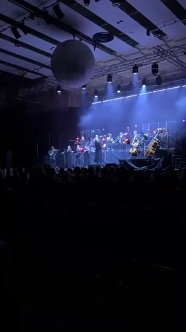 An orchestra performs on a stage in a dark auditorium. The orchestra consists of string, brass, and percussion instruments. A large moon prop hangs above the stage. The audience sits in rows of seats in front of the stage. The stage is lit with blue lights and fog.