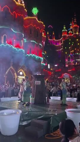 In the video, three individuals in traditional attire are performing on a stage in front of a brightly illuminated building at night. The building, decorated with colorful lights, appears to be a castle or similar structure. A fire burns in a large, primitive-looking brazier at the center of the stage, producing a significant amount of smoke. Two of the performers are positioned on either side of the brazier, while one stands in front of it. Both sides of the stage have large white buckets. A crowd of spectators is visible in the background, gathered to watch the performance. The stage itself is covered in a green tarp, with some fallen flower petals or similar debris scattered around.