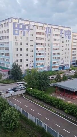 The video shows a view of a residential area. In the foreground, there is a paved area with white markings, resembling a road or a path, bordered by a fence and green bushes. Beyond this, there are two apartment buildings, one taller and lighter colored, the other slightly shorter and a different shade. In front of the buildings, there are trees and parked cars. Further to the right, there is a playground area with structures like slides and a covered seating area. The sky is cloudy. The scene appears to be captured from a slightly elevated perspective, possibly from a window.
