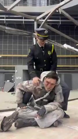 Two men, apparently actors, are goofing around in what appears to be a movie set or studio. One man is dressed in a dark police uniform and the other man is sitting on the floor wearing a military style camouflage outfit that is covered with what looks like dirt and blood. The man on the ground smiles up at the man in the police uniform, who is standing behind him. The background is a large room with a metal framework structure, grid-like walls and scattered debris.