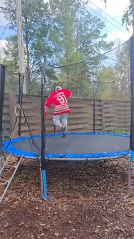 A person in a red and white jersey and grey pants is on a trampoline. They are in a crouched position and then fall onto their stomach. The trampoline is outdoors and surrounded by a wooden fence and trees. The sky is visible and appears to be partly cloudy.