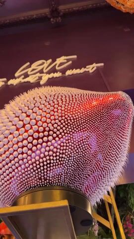 The video features a close-up view of a striking, abstract sculpture with an organic, rounded form. Its surface is densely covered with numerous small, white, conical or spherical protrusions, giving it a highly textured, almost spiky appearance. The interior of the sculpture is illuminated with dynamic, shifting lights, transitioning between vibrant red and purple hues, which glow through the gaps or translucent elements of its structure. The sculpture rests on a golden-colored base or stand.

In the background, a dark-colored wall is visible, adorned with white neon text. The word "LOVE" is clearly discernible in the neon sign, followed by partially visible text that appears to read "what you want". Above and to the right of the sculpture, a large, ornate chandelier or light fixture, composed of numerous orange or gold-colored elements, hangs from the ceiling. The lighting suggests an indoor setting, likely an evening or nighttime ambiance, possibly within a restaurant, bar, or art space in Amersfoort, Netherlands. The video primarily shows a static view of the sculpture with slight camera movement, highlighting its unique design and changing internal illumination.