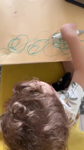In a top-down view, a young child with curly brown hair is seated on a yellow chair or cushion, diligently drawing on a large piece of brown paper. The child holds a blue marker in their right hand, creating abstract scribbles, loops, and circular patterns across the paper. They are wearing a white short-sleeved shirt adorned with a light brown leafy or animal print, and dark socks with grippers. The setting appears to be indoors, possibly at a table, in Pétange, Luxembourg. The natural lighting suggests it's daytime.