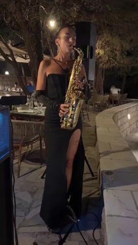 A woman plays the saxophone at an outdoor restaurant at night. She wears a black, strapless, floor-length gown with a high slit on the left leg and long black gloves. Her hair is pulled back, and she wears dangling earrings. The saxophone is golden brass.

The setting is an outdoor dining area with tables and chairs. In the background, trees and other diners are visible. A speaker is set up to her left, and a stone wall with a water feature is to her right. The lighting is dim, suggesting evening or nighttime. The overall atmosphere appears to be one of a live performance at a restaurant.