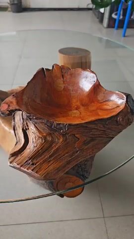 A person's hand wearing a grey sweater is touching a wooden object on a glass table. The wooden object appears to be a decorative bowl carved from a tree stump, with an uneven, natural shape and a glossy finish. The background shows a room with a tiled floor and furniture, including a wooden stool and plants.