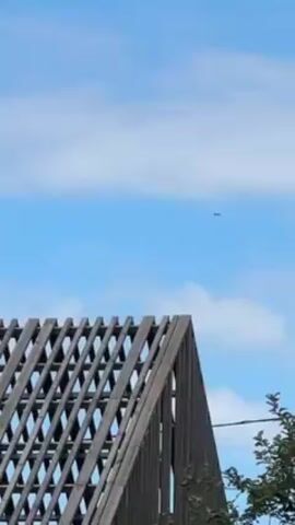 The video shows the top of a dark gray roof structure under a bright blue sky with a few scattered clouds. The roof is made of wooden beams arranged in a lattice pattern. In the distance, a small dark airplane is flying across the sky. A power line and some foliage are visible on the right side of the frame.