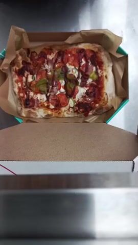The video shows a rectangular pizza in a cardboard box. The pizza is topped with tomato sauce, cheese, pepperoni, and green peppers. A person's hands are visible holding the pizza box and using a knife to cut the pizza. The background appears to be a kitchen or food preparation area with a metal surface. The video focuses on the act of cutting the pizza into slices.
