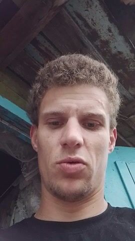 A close-up, slightly blurry, POV video shows a man’s face from a low angle, with his head tilted. He has curly, light brown hair and a light beard. His eyes are wide open and looking directly at the camera. He is wearing a black t-shirt. The background is out of focus and appears to be an outdoor or rustic setting with wooden beams and possibly a blue painted surface. The man makes several facial expressions, including a quizzical look, a pursed-lipped expression, and a smile. The lighting is natural and seems to be coming from above. The video is very short, lasting only a few seconds.