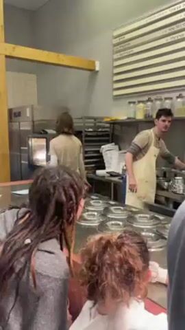 The video captures an indoor scene in what appears to be a restaurant or food service establishment. Several people are present, including a man wearing an apron who seems to be a staff member, busy preparing or serving food behind a counter. The counter has multiple circular containers, some with lids on them. Customers are seen from the front, some waiting at the counter, including one with dreadlocks and another with curly red hair. Behind the staff member is a stainless-steel oven, open with something inside, and a rack full of baking trays. A menu is displayed on the wall behind the counter. The environment seems functional, with food service equipment and storage. The lighting is subdued.