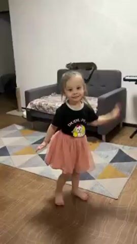 A little girl is dancing in a living room. She is wearing a black t-shirt with a cartoon on it and a light pink tutu. She has her hair up in a ponytail and is barefoot. She is twirling and smiling. A gray sofa with a patterned blanket is behind her and a cat is resting on the sofa. There is a geometric rug on the floor and wooden floorboards. A TV stand is to the left.