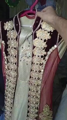 A person's hand is holding a hanger with a decorative garment on it. The garment appears to be a traditional or formal dress, possibly a kaftan or a similar outfit. It is a sleeveless, floor-length dress, made of a light-colored, shimmery fabric. Over this base dress, there is a darker, burgundy-colored overlay that resembles a jacket or a vest. This overlay is heavily embellished with intricate gold embroidery, beads, and possibly sequins, forming floral patterns and borders. The sleeves of the overlay are short and appear to be made of a sheer, reddish fabric, also adorned with gold embellishments. The neckline of the overlay is high and structured, with decorative trim. The garment is displayed against a background that is out of focus but suggests an indoor setting, possibly a closet or a dressing area, with other items of clothing or fabric visible. The lighting appears to be artificial.