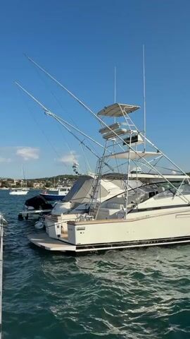 The video shows a large white fishing boat docked in clear blue water under a bright blue sky. The boat has a tall tower structure with multiple levels, suggesting it's equipped for offshore fishing. Fishing rods extend from the top of the tower. The boat has a sleek design with a dark stripe along its hull. Other boats and a distant coastline with buildings and vegetation are visible in the background. The water is choppy with visible waves.