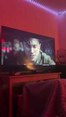 A close-up shot shows a television screen displaying a scene from a Harry Potter movie. On screen, Harry Potter, wearing his signature round glasses, is looking intently at someone off-screen. The subtitles read, "Look at me." The scene then cuts to a close-up of Severus Snape. The TV sits on a light-colored wooden table, covered partially by a fuzzy, pink blanket. The room is illuminated by a pinkish-red LED strip light running along the top of the wall. A black wire basket sits on a dark piece of furniture in the background.