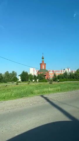 The video shows a side view of a building with an orange and white facade, likely a school or administrative complex, seen from a moving vehicle. In front of the building, there's a small red church with a green dome and a gold cross. A green grassy field is in the foreground, with a dirt road or path. Several trees are scattered throughout the scene, and power lines are visible against a clear blue sky. A billboard with text and images is also present on the right side of the frame. The camera is likely mounted inside a vehicle, capturing the passing scenery.