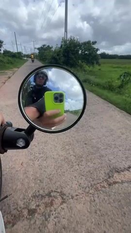 The video shows a person riding a vintage blue and white Honda motorcycle on a narrow paved road. The camera angle is from the driver's perspective, showing the handlebars, speedometer, and the road ahead. Another motorcyclist can be seen in the distance. The driver checks their reflection in the side mirror, revealing a person wearing a helmet and sunglasses holding a bright green smartphone. The environment consists of green vegetation on either side of the road, utility poles, and an overcast sky. The road surface appears to be somewhat worn. The motorcycle’s speedometer shows it’s only gone 6167 kilometers.