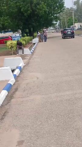 The video shows a gray asphalt road bordered on one side by a curb with a blue and white painted edge. A man in dark clothing is sitting on a concrete block by the curb, looking at something in his hands. Further down the road, three people are walking together. A black SUV is driving away in the distance. On the left side of the road, there is a small green space with some bushes and a red car parked in the background. A street sign and several tall, thin streetlights can be seen.