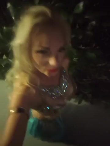 The media file shows a close-up selfie of a woman with blonde, wavy hair and bright red lipstick. She is wearing a bejeweled, ornate costume that appears to be a belly dancing outfit, with a decorated bra and a necklace. The lighting suggests it is nighttime, possibly outdoors, as there are dark shadows and hints of foliage in the background. The woman is looking directly at the camera with a confident expression. The setting appears to be a dimly lit outdoor area, possibly a garden or courtyard. There is no visible text in the media file. The overall mood is glamorous and celebratory.