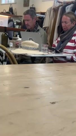 A man and a woman are seated at a wooden table in what appears to be a cafe or shop. The man, wearing a plaid shirt and a khaki jacket, is looking at a laptop and occasionally touching his face. The woman, in a red and white striped shirt and a scarf, is also looking towards the man's laptop. Between them is a straw hat and glasses of water. In the background, there are clothes hanging on a rack and shelves with various items. A bottle of wine is visible on a counter to the left. The overall lighting suggests an indoor setting during the day.