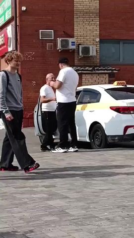 The video shows an outdoor scene in front of a brick building. In the foreground, two young men, one with curly blonde hair and the other with dark hair wearing a blue backpack, walk from left to right. Behind them, an older man in a white shirt and dark pants stands near a white taxi with a yellow stripe. Another older man in a white t-shirt and black pants is also present. The background shows a sidewalk, a flower pot, a street with cars, and apartment buildings under a cloudy sky. The camera pans slightly to the left, revealing more of the street and buildings. In the middle ground, a woman with a cane walks from right to left. A man is seen pulling a suitcase towards the foreground. The video also shows a woman sitting on a bench in the foreground, wearing a dark blue baseball cap and a checkered shirt, with a backpack next to her. She appears to be looking at something in her hands.