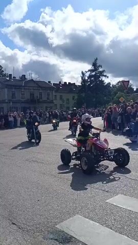 The video shows a parade or event on a paved road. A young person in a helmet and protective gear is riding a red and yellow ATV in the foreground. Behind the ATV, there are several motorcycles, some with riders. On either side of the road, there are crowds of spectators watching the event. Buildings and trees are visible in the background, and the sky is partly cloudy with blue sky. The overall scene suggests a community event or celebration involving vehicles.