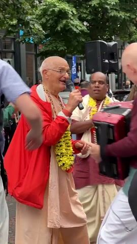 The video shows a man in a red jacket and light-colored clothing playing an accordion and singing into a microphone. He is wearing a garland around his neck. To his right, another person with a shaved head, wearing a maroon shirt and a green vest, is facing away from the camera. To the left of the accordion player, another man in a green jacket and yellow shirt, also wearing a garland, is holding what appear to be small cymbals. The scene appears to be outdoors, possibly a street, with trees and buildings in the background. There are other people visible in the background, but they are out of focus. The overall impression is of a public performance or celebration.