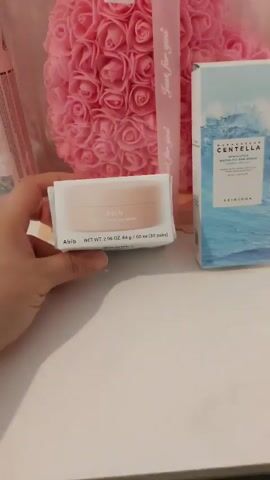 The video displays several skincare products on a white surface, set against a background featuring a large pink floral arrangement made of artificial roses.

In the foreground, a hand holds a white box for "Abib gummy sheet mask eye patch." The box is primarily white with light pink accents and features the brand name "Abib," the product type "gummy sheet mask eye patch," and net weight information: "NET WT. 2.96 OZ. 84 g / 60 ea (30 pairs)." The hand slightly adjusts the box, allowing for a clearer view.

To the right of the hand-held product, a taller, blue and white box stands upright. This box is for "MADAGASCAR CENTELLA WATER-FIT SUN SERUM" by "SKIN1004."

The background is dominated by a large, round arrangement of numerous pink artificial roses. A pink ribbon, tied around this floral arrangement, is visible, with the words "Just for you" printed on it multiple times. A portion of another pink product package is partially visible on the far left of the frame. The setting appears to be indoors, likely a display or vanity area.