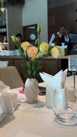 The video showcases a restaurant setting with a focus on a table centerpiece consisting of a vase with a flower arrangement, napkins, and glasses. The flowers are a mix of peach and yellow hues, arranged in a white, textured vase. The table is covered with a white tablecloth, and next to the vase is a holder filled with white napkins folded into a point. An empty water glass sits beside the napkins. In the background, restaurant staff are seen working, including a waiter in a white shirt and brown apron, and another employee in a black cap. There's activity around a counter with stacks of plates, suggesting meal preparation or service. A QR code is visible on a small sign on the table. Overall, the scene presents a well-lit, upscale dining environment.