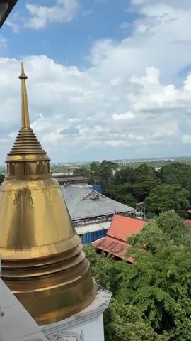 This is a vertical video, panning across a landscape from a high vantage point. In the foreground, on the left, is the golden top of a stupa, with intricate detailing on its tiered structure. The stupa is set against a bright blue sky filled with scattered white clouds.

As the camera pans to the right and slightly down, various buildings become visible. There are several structures with red tile roofs, some nestled among lush green trees. One building appears to be a long, single-story structure with a dark red roof and numerous windows. Further into the background, there are more dense green trees, suggesting a forest or parkland.

Beyond the immediate greenery, the landscape stretches out to reveal a more distant view. A highway or bridge structure is visible crossing a body of water or flat terrain. In the far distance, a city skyline with numerous high-rise buildings can be seen against the horizon.

The overall impression is one of a serene, perhaps spiritual, location offering panoramic views of both natural and man-made environments. The lighting suggests it is daytime, likely midday or early afternoon, with clear, bright sunlight. There are no people or animals visible in the frame.