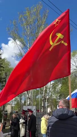 The video shows a gathering of people around a war memorial on a bright day. A large red flag with a gold hammer and sickle is prominently displayed, along with Russian flags and smaller red flags with Cyrillic text. Many people are dressed in civilian clothes, while some wear military-style uniforms with hats. People are standing, walking, and holding pictures or portraits. A tall grey obelisk with Cyrillic inscriptions stands next to a statue of a soldier. Trees are visible in the background under a blue sky with scattered clouds.