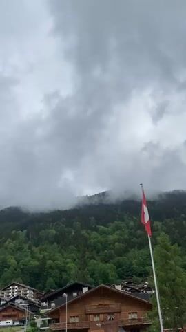 The video captures a time-lapse of a mountainous village in Champéry, Switzerland, under a cloudy sky. In the foreground, traditional wooden chalets are visible. A prominent red flag with a white cross, identifying it as the Swiss flag, stands on a pole on the right side of the frame. Behind the village, a densely forested mountain rises, its upper slopes shrouded in thick, moving clouds. As the video progresses, a patch of blue sky briefly appears through the clouds on the left. Cables, possibly for a ski lift or power lines, stretch across the scene on the left, leading up the mountain. A red and white road sign, partially visible on the left, shows a white bicycle symbol on a blue circle.