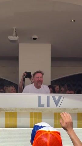 Here is a detailed description of the video:
The video shows a section of a nightclub or beach club, specifically a raised area or VIP section with a white ledge that reads "LIV Beach" in large letters. Below the ledge, there is a yellow and white tiled wall. Behind the ledge, there are several people, including a man in a white t-shirt who appears to be the focus of the video. Someone behind him is holding a camera, possibly taking photos or videos. The man is looking towards the camera with a slight smile. The crowd behind the ledge seems to be enjoying the music. In the foreground, a person is wearing an orange and blue baseball cap. The ceiling has a projector and a security camera. A large screen above displays changing patterns, possibly visuals associated with the music. The overall atmosphere appears lively and energetic.