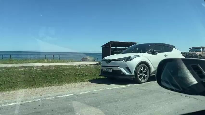 The video shows a traffic scene on a sunny day, likely near a coast, with several cars parked on the side of a road and others driving. The first car in view is a white SUV. Further down the road, a silver SUV and a black SUV are parked. Beyond them, more cars are visible, including a light-colored SUV with a roof rack. The road appears to be paved, and there is a grassy area and a body of water in the background, suggesting a coastal environment. The sky is clear and blue. The perspective suggests the video is filmed from inside a moving vehicle.