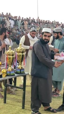This video shows a man in a dark robe and a white kufi shaking hands with a person in a yellow shirt, who appears to be a referee, as the referee holds some papers. To the left, two large golden trophies are displayed on a table. In the background, a crowd of people is gathered, and the event appears to be outdoors on a grassy field. The atmosphere seems celebratory.