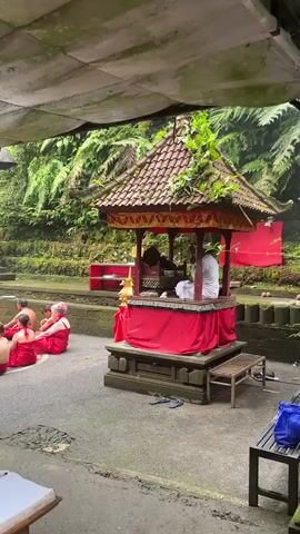 A group of people in red sarongs are sitting on the ground in front of a small shrine. A person in white is standing at the shrine, seemingly performing a ritual. The shrine is decorated with red cloth and has a tiled roof. In the background, there are lush green plants and trees, suggesting an outdoor or semi-outdoor setting. The ground appears to be made of concrete or stone. There is a canopy overhead, possibly for shade. The overall atmosphere seems solemn and spiritual.