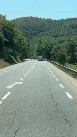 A white van drives down a paved road flanked by green trees and rolling hills under a clear blue sky. The road has white dashed lines marking the lanes and a white arrow indicating a turn. A guardrail runs along the right side of the road.