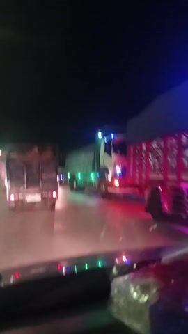 A dashcam view at night shows a line of trucks on a wet road. The camera is inside a vehicle, with a dashboard and parts of the interior visible in the foreground. The truck directly ahead and to the right is a white and blue semi-truck with green and blue lights on its side and front. Its brake lights are illuminated red. To the left and further ahead, another truck is partially visible, its rear cargo area filled with wooden crates. The road surface reflects the lights of the trucks and potentially streetlights. The sky is dark, indicating nighttime. The traffic appears to be moving slowly or stopped.