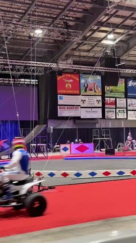 The video shows a circus act involving a motorcycle flying through the air. The environment is an indoor arena or large event space with a high ceiling and multiple sponsor banners displayed on the wall. In the foreground is a circus ring with red carpeting and a diamond-patterned border. An inflatable landing pad with "406 FMX" printed on it is positioned behind the ring. Wires and rigging are visible above the landing pad, presumably used for the motorcycle stunt. At one point, a man in a blue suit and a red fez hat walks into frame, likely a circus performer or organizer. The sounds of the motorcycle and music are audible.
