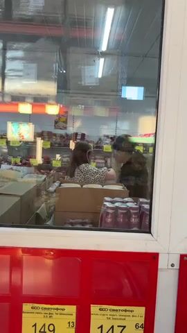The video shows an interior view of a store, filmed through a glass panel. In the background, shelves are stocked with various packaged food items, displaying price tags. In the midground, cardboard boxes are stacked. Two women are visible through the glass. The woman on the left has long, light brown hair and wears a light-colored, animal-print top, facing away from the camera. The woman on the right appears older, with her dark hair covered by a patterned scarf in brown and earthy tones, wearing a dark jacket. At the beginning, the two women are in close proximity, with the older woman leaning towards the younger woman's head. The older woman then turns to her right, faces the shelves, and begins selecting items, placing them into a colorful, floral-patterned reusable shopping bag. The younger woman remains relatively stationary, looking at items on a counter or shelf.