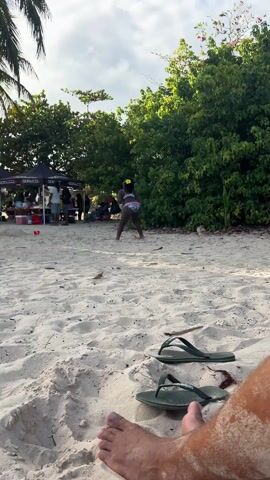 Here's a description of the video:

The video is shot from a low angle on a sandy beach. In the foreground are a pair of green flip-flops and the bare feet of the person holding the camera.  A young, dark-skinned girl in a swimsuit dances barefoot in the sand, moving away from the camera. Behind her, to the left, is a black tent with people underneath it. In the background, lush green trees create a natural backdrop. The sky is overcast.