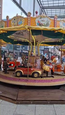 A carousel turns in an outdoor covered area. The carousel has a yellow canopy with green trim, decorated with images of children and various scenes. Lights are strung along the canopy's edge. Various rideable objects are attached to the carousel, including a red car, a green dinosaur, a fire truck with the number 112 on it, and a white bicycle. Several people are visible, some appearing to be children riding the carousel, and others standing around. The background shows buildings and a street scene. The overall impression is that of a lively, public space, possibly a market or town square.