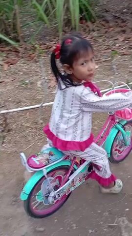 A young girl with dark hair in pigtails, wearing pajamas with a white and pink striped pattern, rides a pink and teal bicycle. The bicycle has a pink basket on the handlebars and decorative decals on the frame. She is on a dirt path outdoors, with green foliage and trees surrounding her. The path appears to lead through a residential area, as a building with a window and a decorative fence are visible to the right. The girl's face shows an expression of joy and concentration as she pedals. The video is a short, continuous recording of her riding the bicycle.