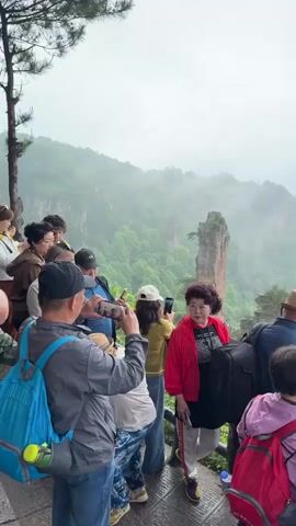 A group of people, mostly older adults, are standing on an observation deck taking photos with their phones. They are looking out at a foggy mountain landscape with tall, rocky pillars covered in green vegetation. The sky is overcast. Some of the people are wearing backpacks. The deck has a metal railing.