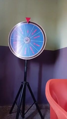 The video shows a standing prize wheel. The wheel is round, blue, and divided into sections, some labeled "TRY AGAIN" and others labeled "FACE CAP" or "BUCKET CAP". It is mounted on a black tripod stand. A red pointer is positioned at the top of the wheel. To the right of the wheel is an orange plastic container, possibly a tub or a chair. The background consists of a wall painted in two colors: a light green upper portion and a dark purple lower portion. The floor is a speckled gray.