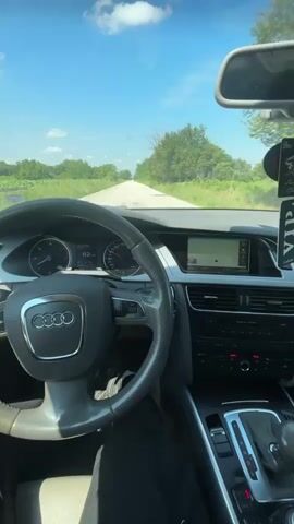 The video captures a first-person perspective from the driver's seat of an Audi car, driving along a rural road on a bright, sunny day.

The steering wheel, adorned with the Audi logo, is prominently featured in the foreground. The dashboard displays a digital speedometer showing the car's speed fluctuating between 82 km/h and 83 km/h. Other visible gauges include the tachometer, fuel gauge, and temperature gauge. A central infotainment screen shows a map or navigation interface. The gear shifter for the automatic transmission is visible on the right, set to 'D' for drive. Hanging from the rearview mirror is a black air freshener or charm with the letters 'VIP' clearly visible. The car's digital clock displays "13:30".

Outside the car, a narrow asphalt road stretches ahead, flanked by dense green trees and bushes on both sides. The sky is clear blue with scattered white clouds, indicating sunny weather during the daytime. The car appears to be traveling at a steady, moderate speed through the countryside, specifically in Brenitsa, Bulgaria, as indicated by the location context. No other vehicles or people are visible on the road.