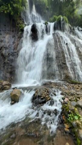 This is a video of a cascading waterfall in a lush, green environment. Water flows down a rocky incline, creating multiple streams and turbulent pools at the base. Trees and vegetation frame the scene, with the sky visible above. The focus is on the natural beauty and movement of the water.