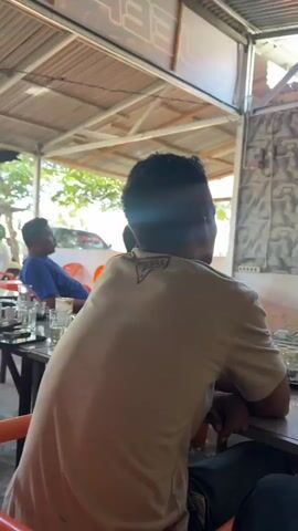 The video shows a group of people sitting at tables, presumably in an outdoor seating area. A television screen is visible in the background, displaying what appears to be a soccer match. The camera focuses on a man in the foreground, seen from behind, wearing a tan t-shirt. He is seated at a table with several glasses and what looks like a cigarette pack. The environment appears to be a casual establishment, possibly a cafe or restaurant, with a corrugated metal roof and simple furniture. The lighting suggests it is daytime. The man in the foreground turns his head slightly, and other people are visible in the background, some also looking towards the television. The video is shaky, indicating it was likely recorded by someone holding a mobile device.
