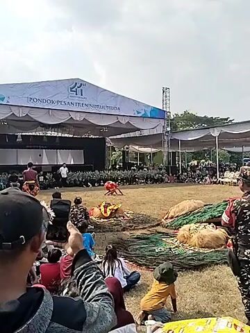 This is a medium shot video of an outdoor cultural performance. In the foreground, the back of a person wearing a black hat and a gray jacket is visible. To their right, other audience members are seated on the grass, including a child in a yellow shirt and a person in a camouflage uniform. In the mid-ground, a performer in a red and yellow costume is engaged in a dynamic dance on a grassy field. Several large, elaborate costumes, possibly for Barongan dancers, are arranged on the ground around the performance area. In the background, a large stage is set up with a white banner that reads "PONDOK PESANTREN NURUL HUDA 41ST ANNIVERSARY." A crowd of spectators is seated in front of the stage, and various stage equipment, including speakers and lighting rigs, are visible. The overall atmosphere appears to be a festive cultural event under a clear sky.