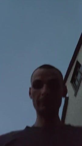 The video shows a man's face from a low angle. He has short hair and a dark t-shirt. He is standing in front of a brick building with a window. The sky is visible in the background. The lighting is dim. The man is looking at the camera. He blinks several times.