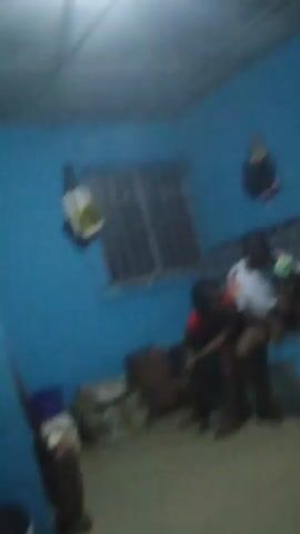 In a dimly lit room with blue walls and a pale ceiling, two people are visible. One individual in a red shirt is seated, while another person in a white shirt with dark shorts sits beside them on a couch or bench. The person in the white shirt appears to be holding something green in their hand. The room contains several bags, one by the window and another near the floor, along with a broom-like object standing in the corner. A window with bars is present, and light is coming from an overhead source. A dark bag also hangs on the wall. The person in the white shirt stands up and walks in place for a moment.